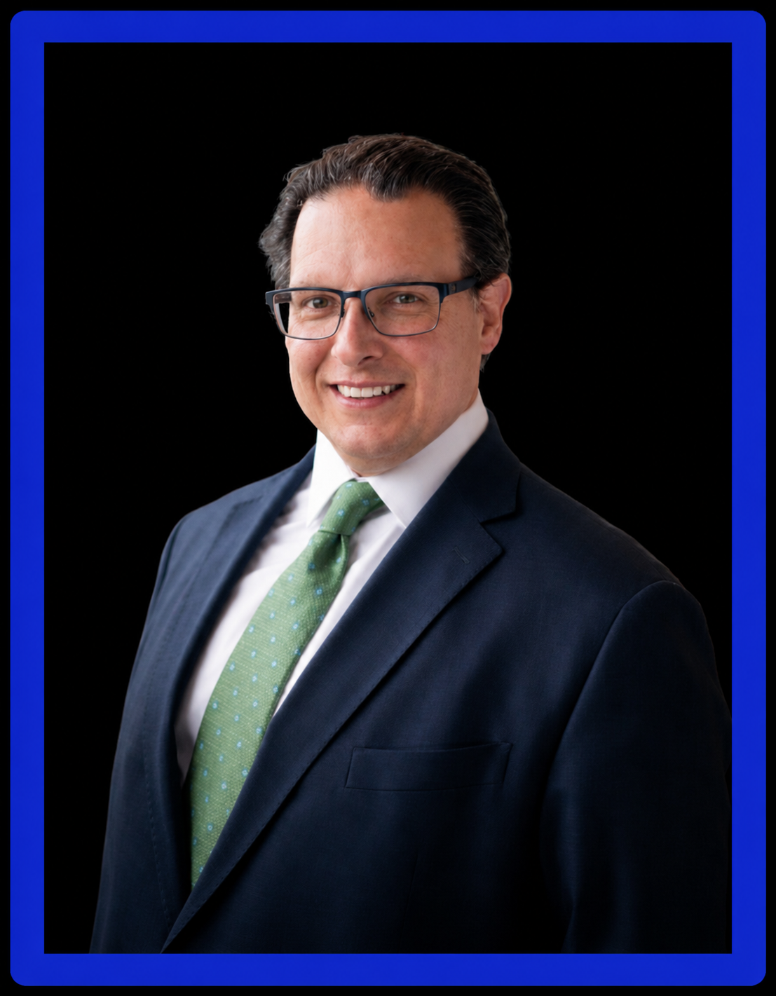 Professional portrait of Cisco Minthorn wearing glasses, a navy suit and light-green tie, against a black background with a royal-blue frame.